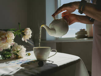 Ältere Frau gießt Tee in Tasse in der Nähe von Blumen zu Hause - IEF00527