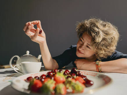 Lächelnde reife Frau, die eine Kirsche vom Teller auf dem Tisch zu Hause aufhebt - IEF00535
