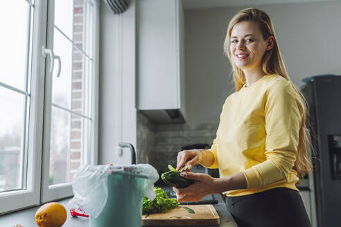 Lächelnde junge Frau beim Schälen von Zucchini am Küchentisch zu Hause - OLRF00153