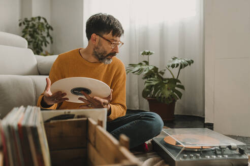 Mann hält Schallplatte und schaut auf den Plattenspieler im Wohnzimmer - DMGF01219