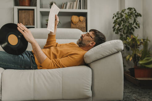 Mann liest Zeitung und liegt mit einer alten Schallplatte auf dem Sofa zu Hause - DMGF01223
