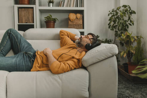 Man listening to music through headphones lying on sofa in living room at home - DMGF01224