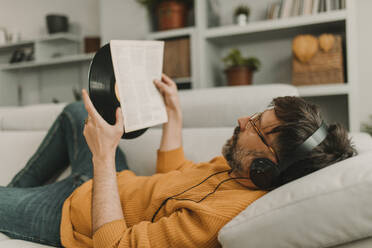 Mann, der über Kopfhörer Musik hört und Zeitung liest, hält zu Hause eine Schallplatte in der Hand - DMGF01226