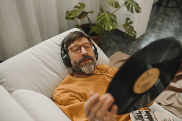 Mann hört Musik und betrachtet eine Schallplatte auf dem Sofa liegend - DMGF01228