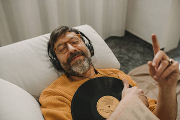 Mann hört Musik über Kopfhörer und liegt mit Schallplatte auf dem Sofa zu Hause - DMGF01229