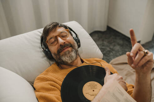 Man listening to music through headphones lying with record on sofa at home - DMGF01229