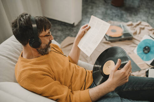 Man listening to music and reading paper holding record at home - DMGF01230