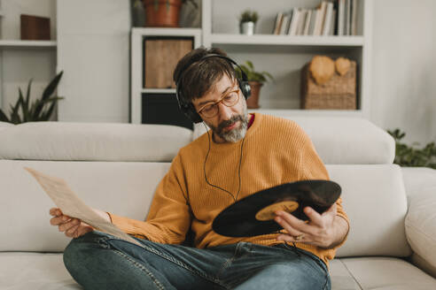 Man listening to music and reading paper holding record sitting on sofa at home - DMGF01231