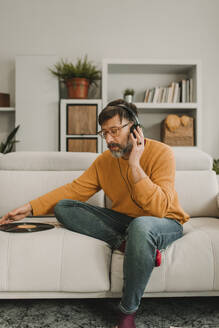 Mann hört Musik über Kopfhörer und sitzt mit Schallplatte auf dem Sofa zu Hause - DMGF01232