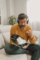 Mann, der zu Hause auf dem Sofa mit einer Schallplatte sitzt und über Kopfhörer Musik hört - DMGF01233