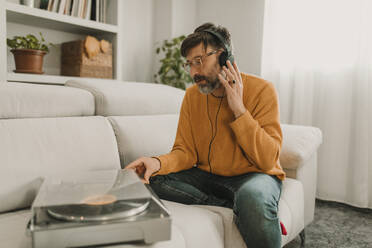 Mann, der zu Hause auf dem Sofa neben einem Plattenspieler sitzt und über Kopfhörer Musik hört - DMGF01234