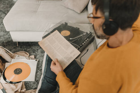 Mann hört Musik über Kopfhörer und liest Zeitung, während er auf dem Sofa zu Hause neben einem Plattenspieler sitzt - DMGF01240