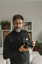 Man holding old-fashioned instant camera standing at home - DMGF01245