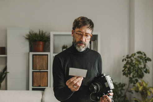 Man holding old instant camera and photograph standing at home - DMGF01250