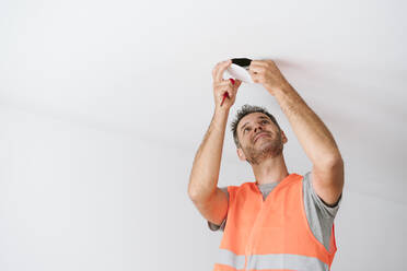 Mature technician installing electric light on ceiling at home - EBBF08634