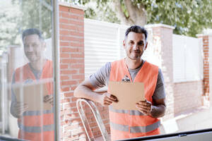 Lächelnder Handwerker steht am Fenster eines Hauses - EBBF08658