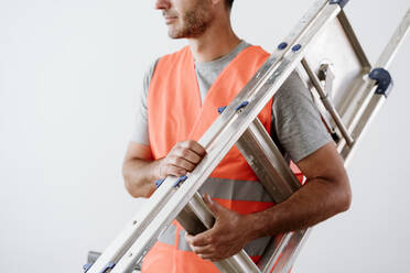 Mature technician carrying ladder in front of wall at home - EBBF08660