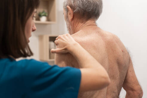 Eine Physiotherapeutin hilft einem älteren Mann während einer Rehabilitationsmaßnahme zu Hause, wobei sie sich auf seinen Rücken konzentriert - ADSF52840
