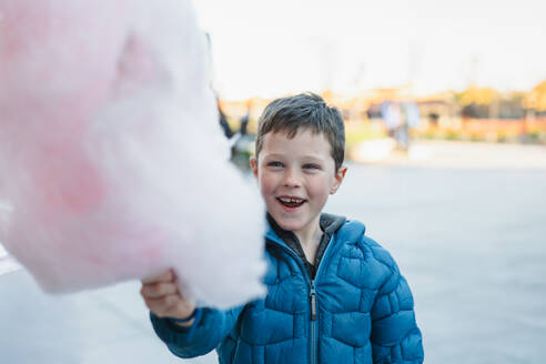 Ein fröhlicher Junge, der eine große rosa Zuckerwatte in einer Umgebung im Freien hält - ADSF52848