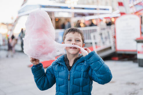 Glücklicher Junge in einem blauen Mantel, der in die Kamera schaut, während er mit einem großen Stück Zuckerwatte auf einem Rummelplatz posiert - ADSF52849