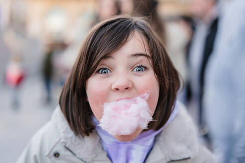 Nahaufnahme eines aufgeregten Mädchens mit großen Augen, das in die Kamera schaut, während es einen Bissen Zuckerwatte auf einem unscharfen Straßenhintergrund genießt - ADSF52850