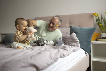 Ein fröhlicher Großvater genießt die Spielzeit mit seinem kleinen Enkelkind, umgeben von Plüschtieren auf einem gemütlichen Bett - ADSF52861