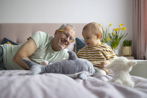 Ein fröhlicher Großvater liegt neben seinem glücklichen kleinen Enkelkind, das mit Stofftieren auf einem gemütlichen Bett spielt - ADSF52863