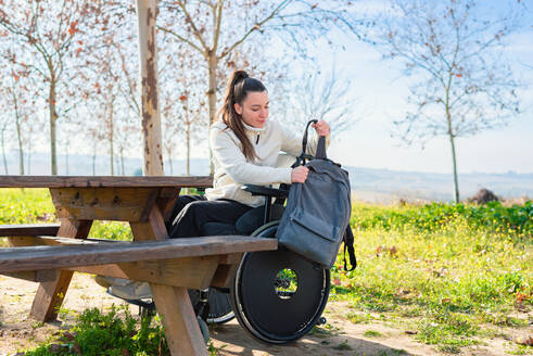 Eine junge Frau im Rollstuhl sortiert ihren Rucksack an einem Tisch im Park - ADSF52871