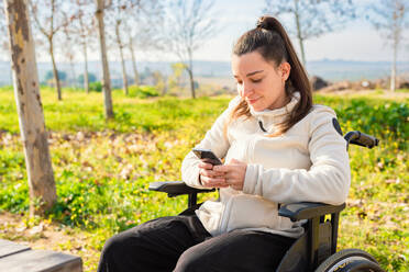 Eine junge Frau im Rollstuhl lächelt, während sie in einem Park auf ihrem Smartphone surft - ADSF52874