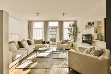 Ein geräumiges, modernes Wohnzimmer, das in natürliches Sonnenlicht getaucht ist, mit bequemen beigen Sofas, Holzfußböden und stilvollem Dekor. - ADSF52919