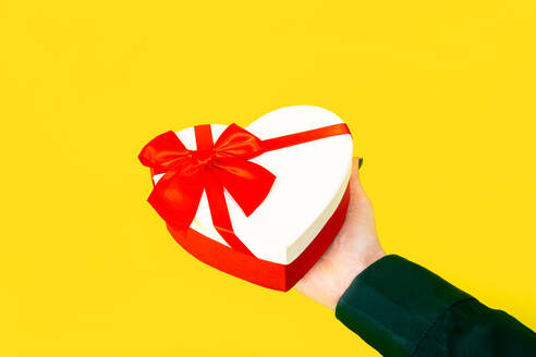 Anonyme Hand, die einen herzförmigen Geschenkkarton mit roter Schleife vor gelbem Hintergrund hält - ADSF52932