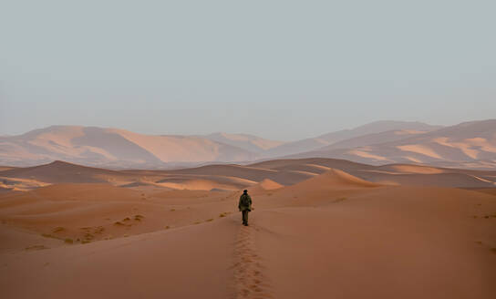 Rückenansicht einer einsamen, nicht erkennbaren Person, die sich auf ein Abenteuer durch die sanft geschwungenen Sanddünen der Wüste von Merzouga begibt, unter den sanften Farben eines marokkanischen Sonnenuntergangs. - ADSF52937