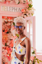 Portrait of cheerful young woman looking at camera in stylish outfit and sunglasses. She is standing near pink phone both and blooming flowers - ADSF52940