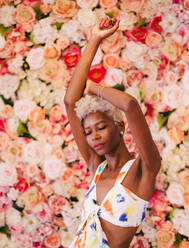 Portrait of sensual young panamanian woman with eyes closed in stylish summer outfit. She is raising arms besides a beautiful blooming pink flowers wall - ADSF52943