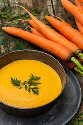 Frische Karottensuppe in einer Schale mit ganzen Karotten auf einem rustikalen Holztisch, garniert mit Petersilie. - ADSF52948