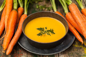 Frische Karottensuppe mit Petersilie in einer schwarzen Schale, Karottenbündel an der Seite, Holzoberfläche. - ADSF52949