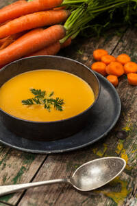 Eine Schale mit cremiger Karottensuppe, garniert mit frischen Kräutern, umgeben von ganzen und geschnittenen Karotten auf einem Holztisch. - ADSF52950