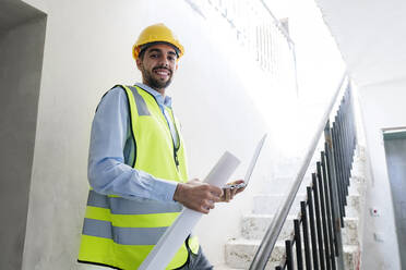 Smiling architect wearing hardhat and standing on steps holding laptop at site - AAZF01496