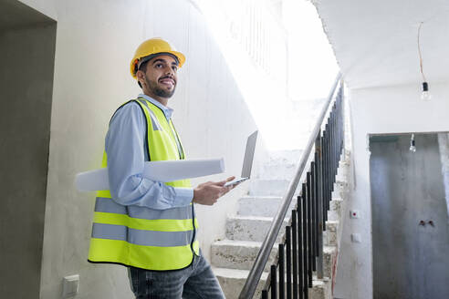 Lächelnder Architekt mit Warnweste und auf den Stufen einer Baustelle stehend - AAZF01498