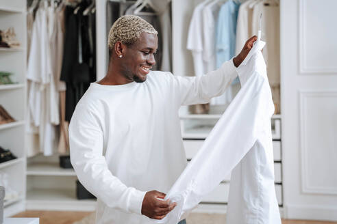 Glücklicher Mann betrachtet ein T-Shirt vor einem Kleiderschrank - NLAF00305