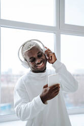 Lächelnder Mann, der drahtlose Kopfhörer trägt und ein Smartphone benutzt - NLAF00310