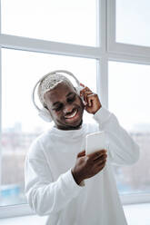 Smiling man wearing wireless headphones and using smart phone - NLAF00310