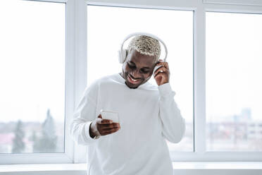 Happy young man wearing wireless headphones and using smart phone - NLAF00311