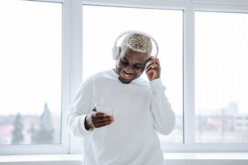 Glücklicher junger Mann, der drahtlose Kopfhörer trägt und sein Smartphone benutzt - NLAF00311