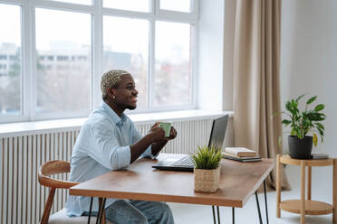 Lächelnder Freiberufler sitzt mit Kaffeetasse und Laptop am Schreibtisch - NLAF00355