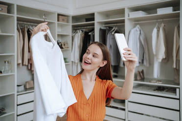 Glückliche junge Frau nimmt Selfie mit T-Shirt durch Smartphone zu Hause - NLAF00370