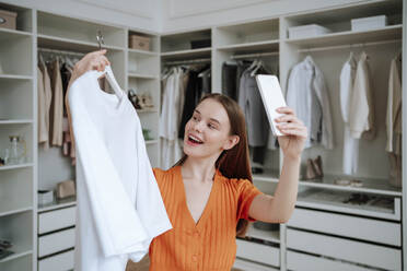 Happy young woman taking selfie with t-shirt through smart phone at home - NLAF00370