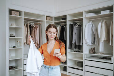 Smiling woman using smart phone near closet - NLAF00371