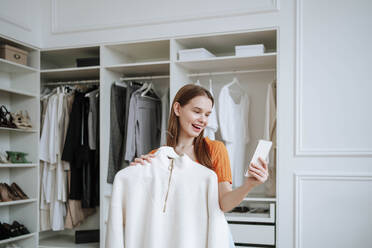 Happy woman talking on video call near closet at home - NLAF00373