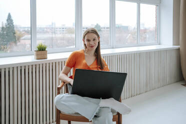 Lächelnder junger Freiberufler, der mit einem Laptop auf einem Sessel zu Hause sitzt - NLAF00374